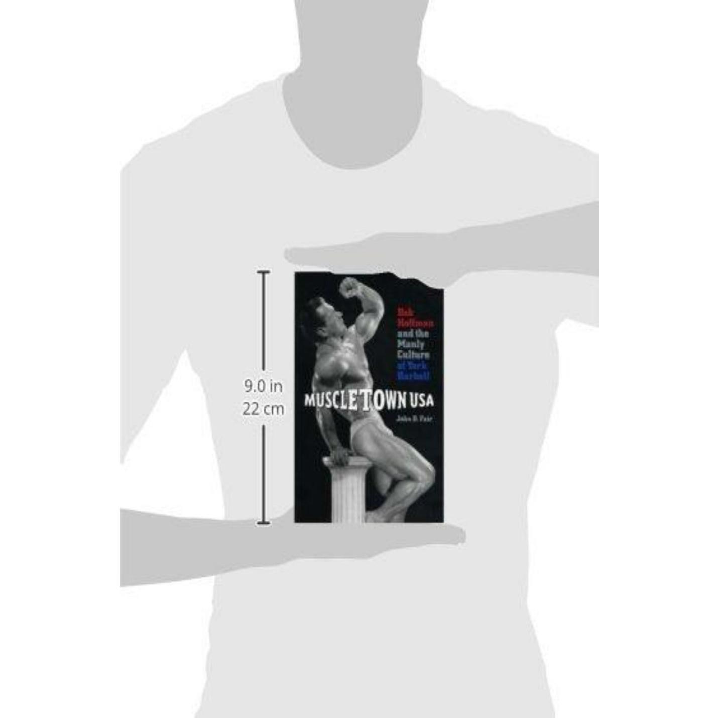 Een grafisch T-shirt met de boekomslag van "John D Fair: Muscletown USA: Bob Hoffman and the Manly Culture of York Barbell" van John D. Fair, met een zwart-wit afbeelding van een bodybuilder die op een voetstuk poseert.