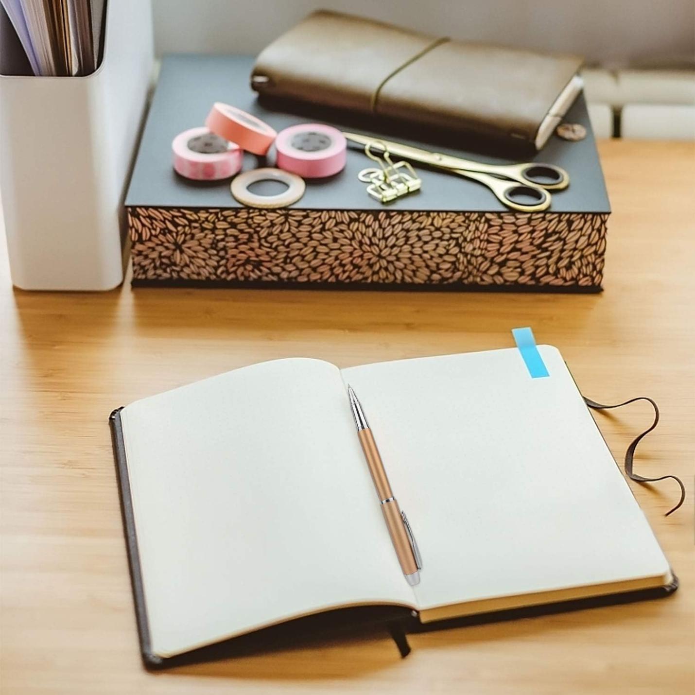 Een netjes ingedeeld bureau met een opengeslagen notitieboekje, een Laat een blijvende indruk achter met deze elegante en hoogwaardige rollerbalpen, een schaar en rollen washi-tape.