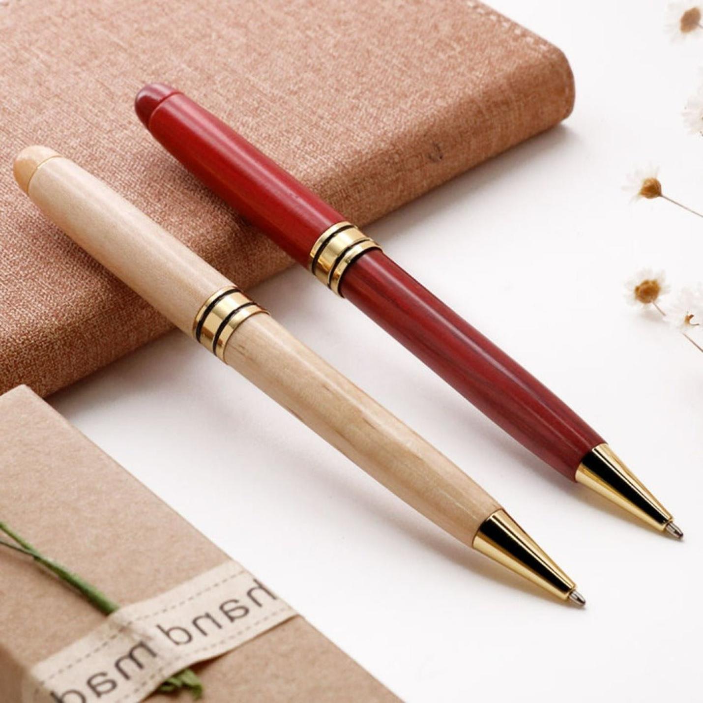 Twee elegante Schrijfsters in stijl en draag bij aan het milieu met onze eco houten balpennen rustend op een stoffen oppervlak met een bloemig accent en ingepakte pakjes op de achtergrond.