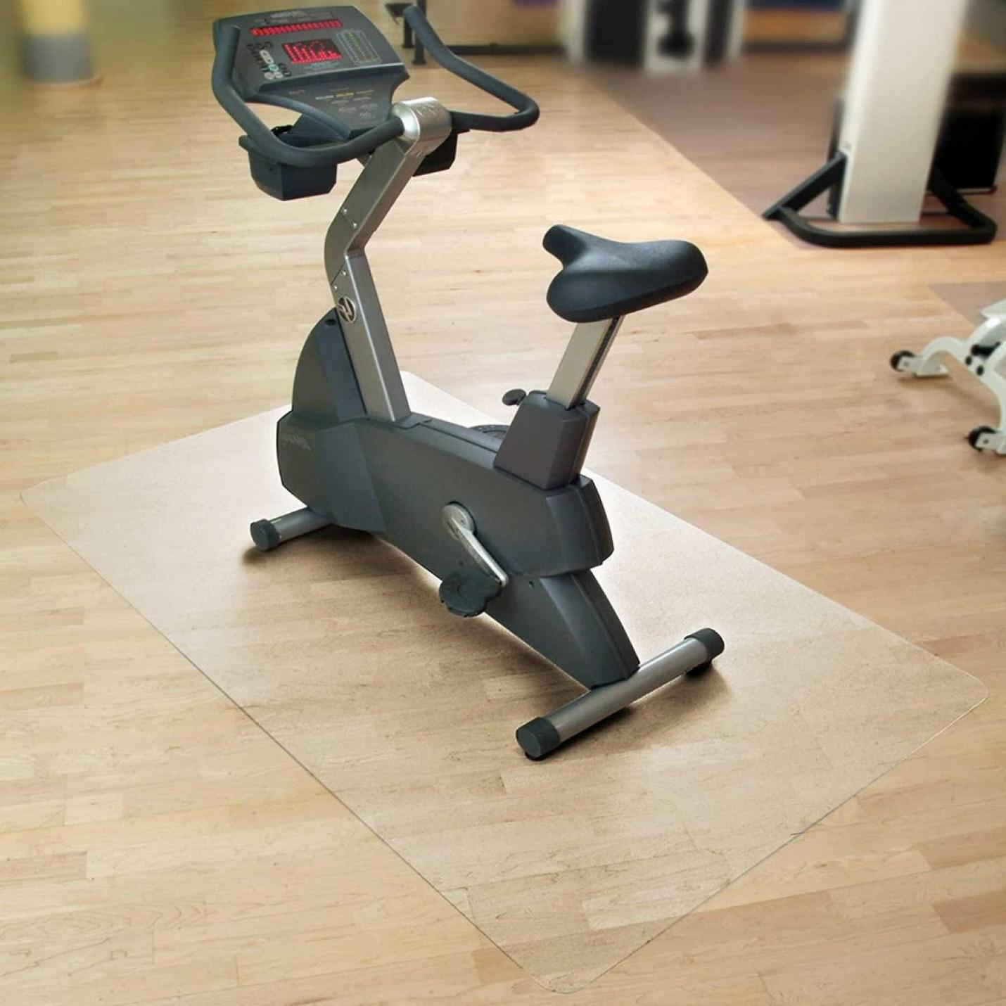 Een hometrainer geplaatst op een Beschermmat voor sporttoestellen boven een houten vloer in een ruimte met fitnessapparatuur op de achtergrond.