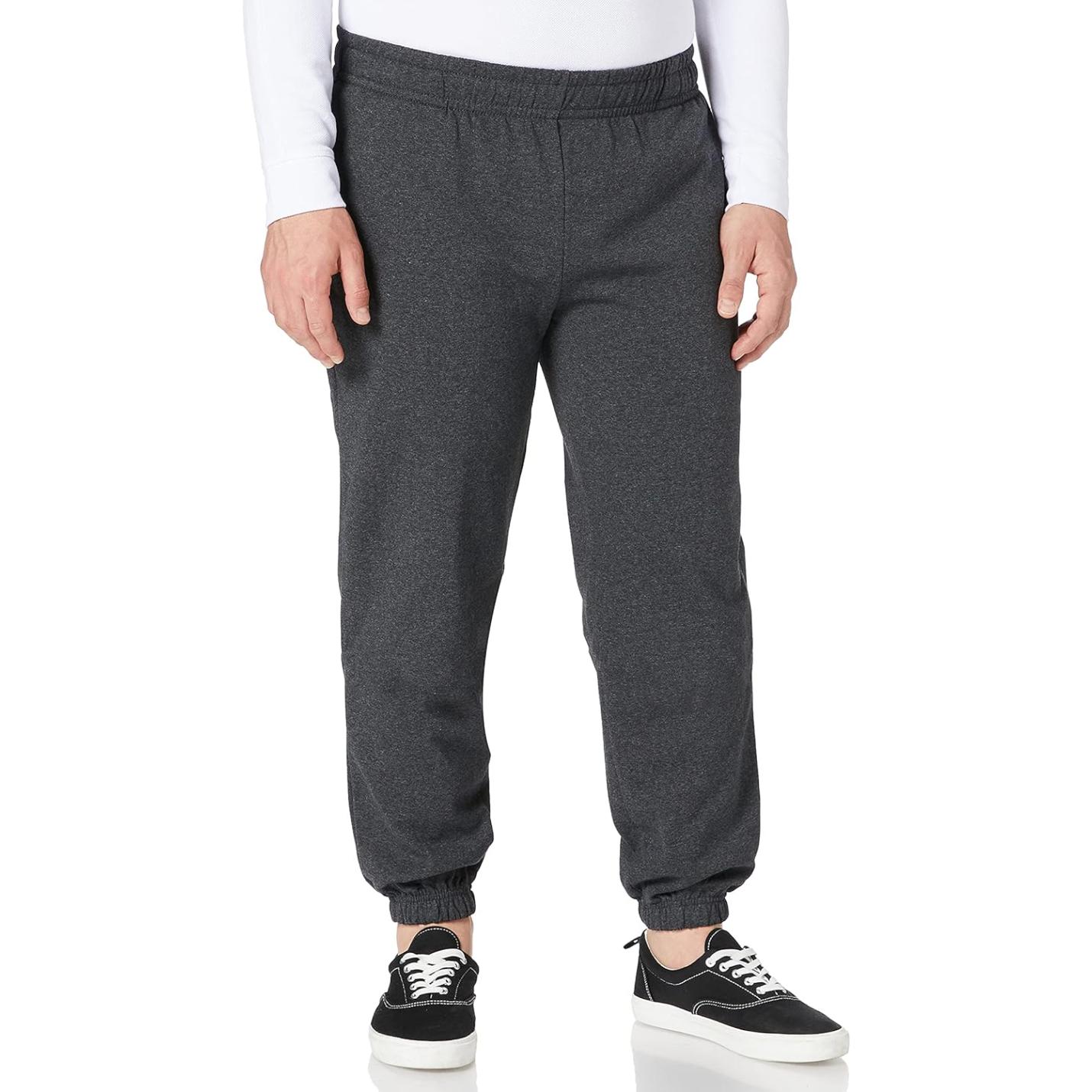 Een persoon die de "🏃‍♂️ Comfort, stijl en bewegingsvrijheid – De ultieme basic joggingbroek!" in een donkergrijze tint, gecombineerd met zwarte sneakers met witte zolen, staat tegen een witte achtergrond.