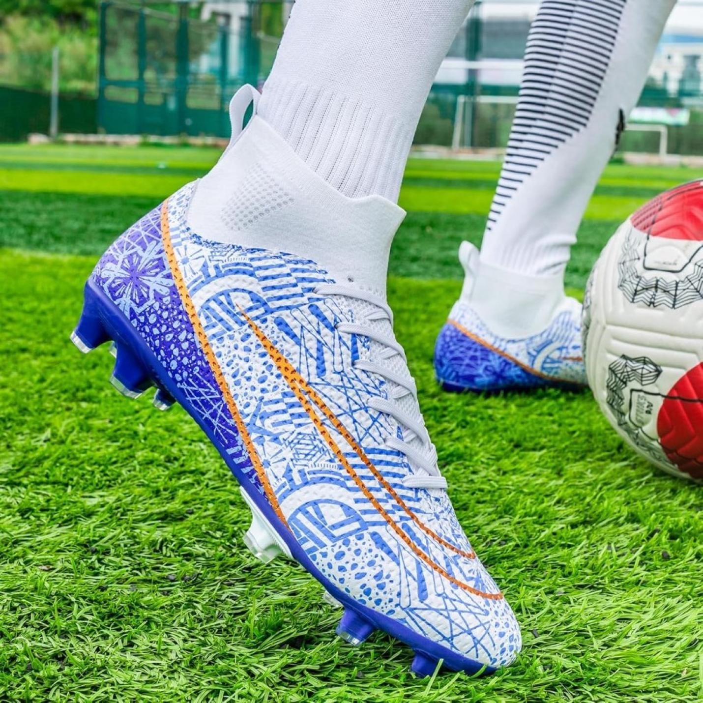 Close-up van een persoon die hoogwaardige voetbalschoenen draagt op een grasveld. Het witte ontwerp met blauwe en oranje patronen en de hoge schoenen voor grip en stabiliteit zijn goed te zien, naast een rood-witte voetbal.