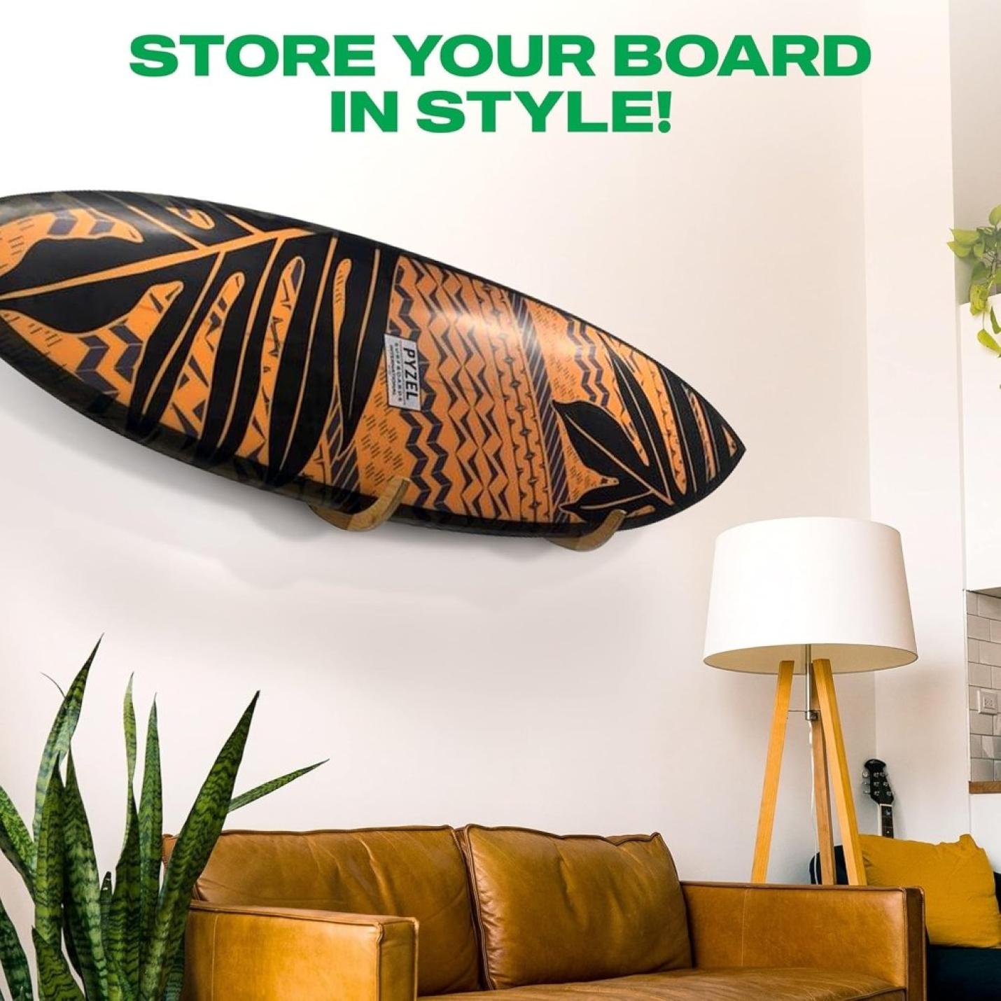 De "Jouw surfplank verdient een stijlvolle display! 🏄‍♂️" hangt aan een witte muur boven een bruine leren bank met een staande lamp en een hoge plant ernaast. De tekst bovenaan luidt: "STORE YOUR BOARD IN STYLE!.