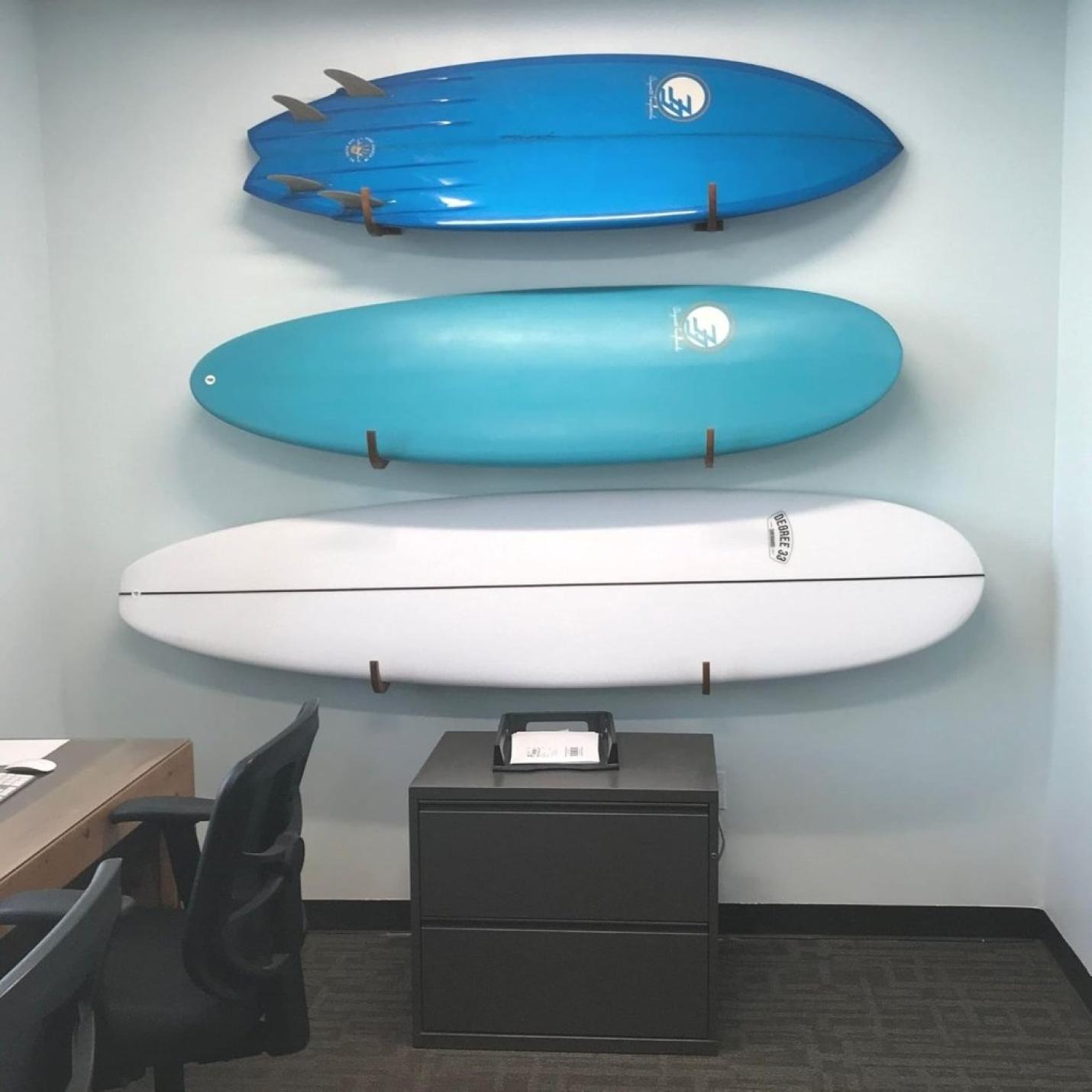 Drie surfplanken in blauw en wit, elegant gemonteerd met de tekst "Jouw surfplank verdient een stijlvolle display! 🏄‍♂️", zweven boven een zwarte archiefkast in een kantoorruimte met een bureau en een stoel.