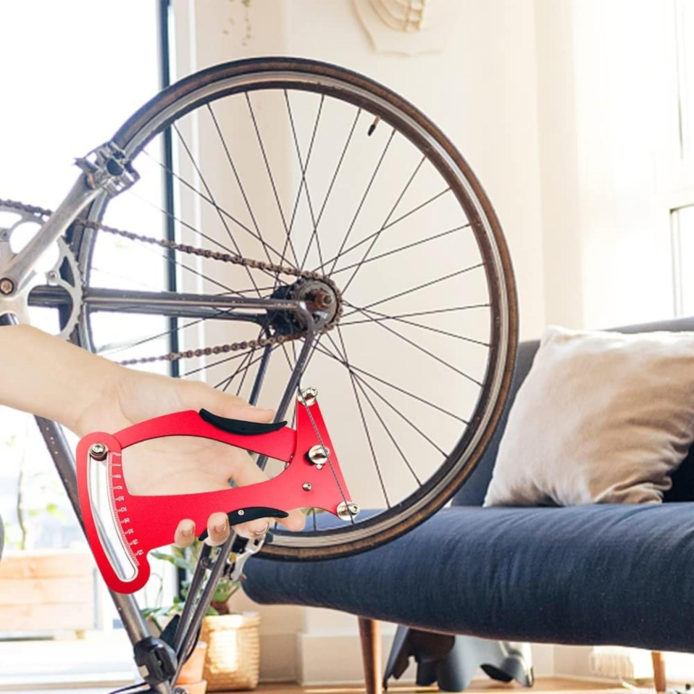 Een hand houdt de 🔧 Ontmoet en optimaliseer je fietsspanning met deze spaakspanningsmeter! vlakbij een omgekeerde fiets binnenshuis met een bank op de achtergrond, klaar om af te stemmen voor optimale wielprestaties.